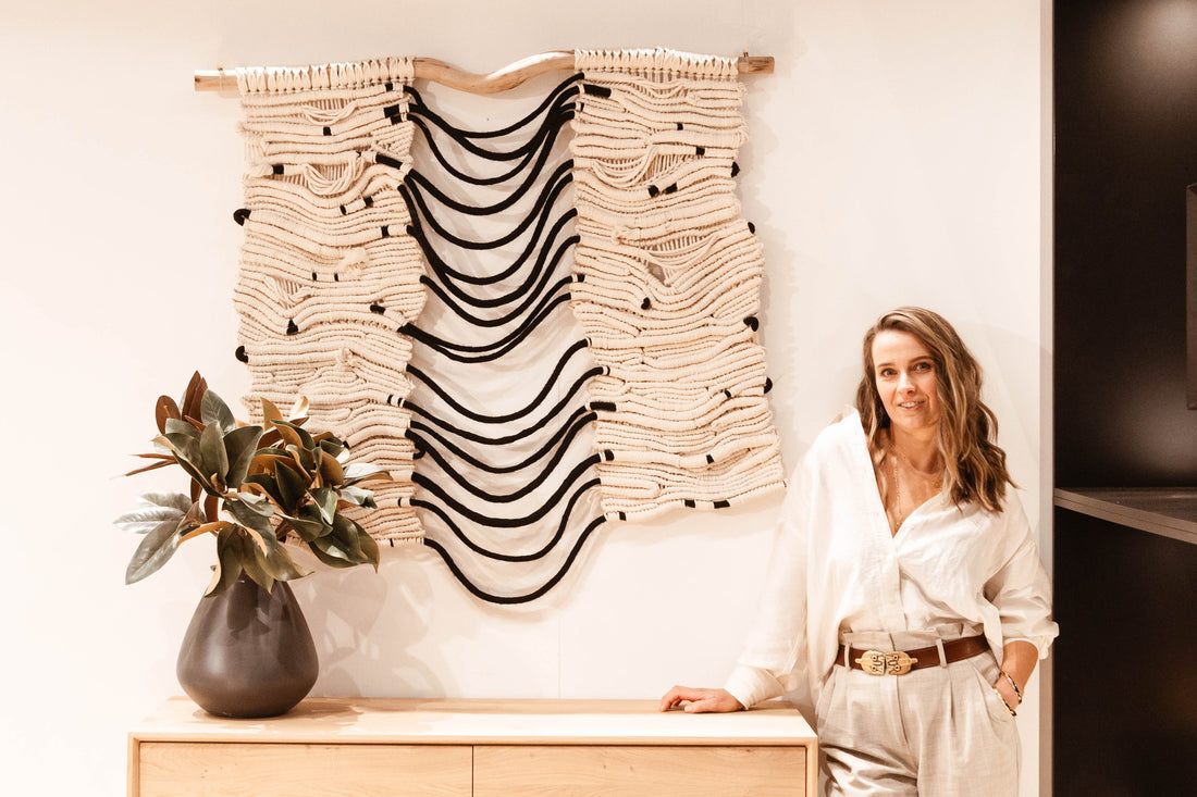 Artist standing next to a custom fibre art installation made of natural materials in neutral tones with black accents, displayed above a light wood sideboard with a black ceramic vase holding greenery.