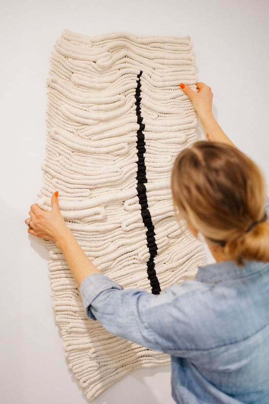 Person arranging a textured, beige fibre art piece on a white wall.